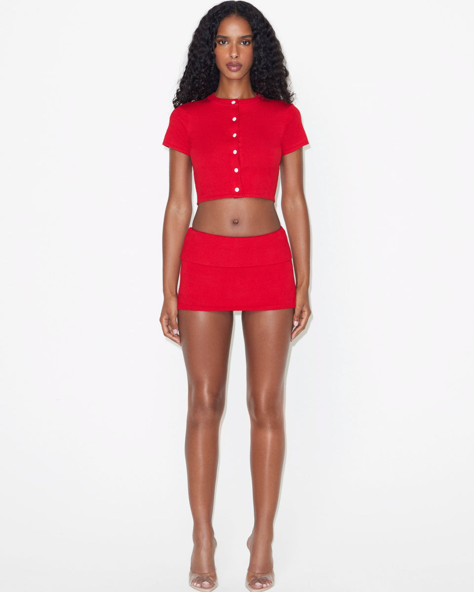 Mini skort in red styled with a short-sleeve buttoned crop top, shown from the front on a model in clear heels.